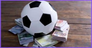 a soccer ball on top of a pile of money in different currencies, on a wooden surface, with a purple border.