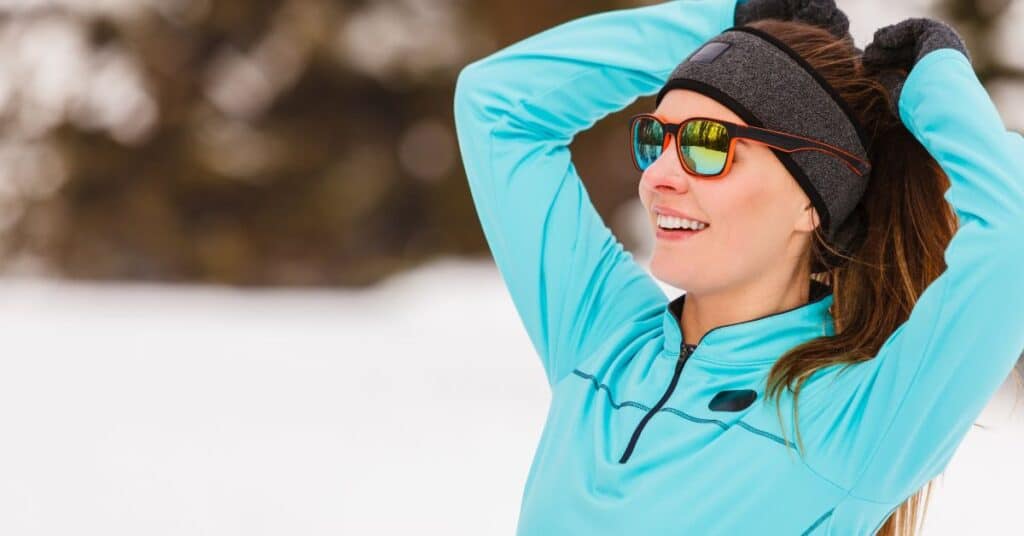 A athletes woman in a blue shirt and a black headband stretching their arms in a snowy landscape.