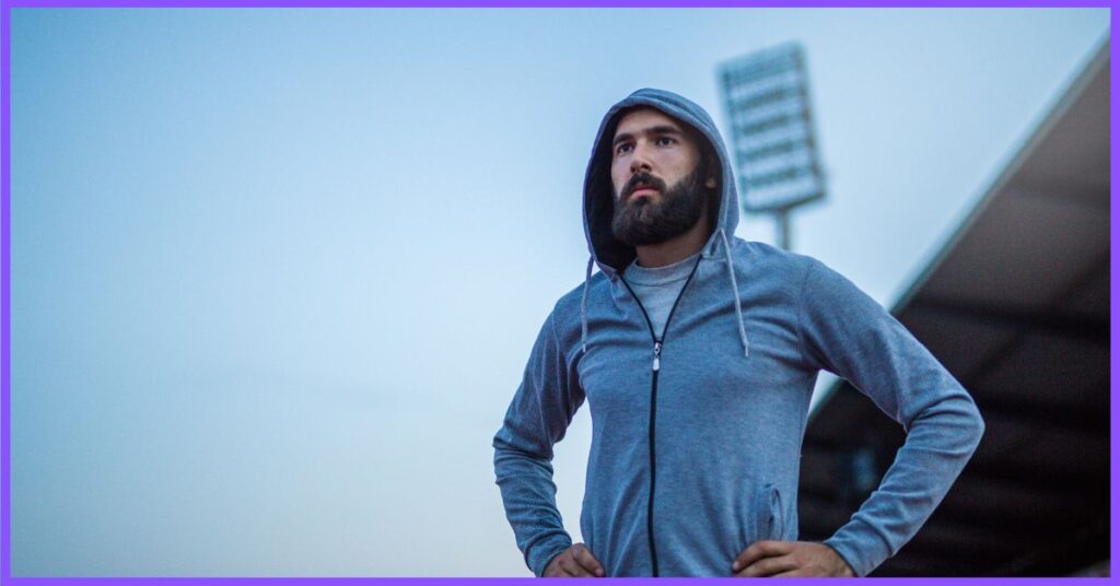 A man in a gray hoodie and earphones standing in front of a stadium with a blue sky.