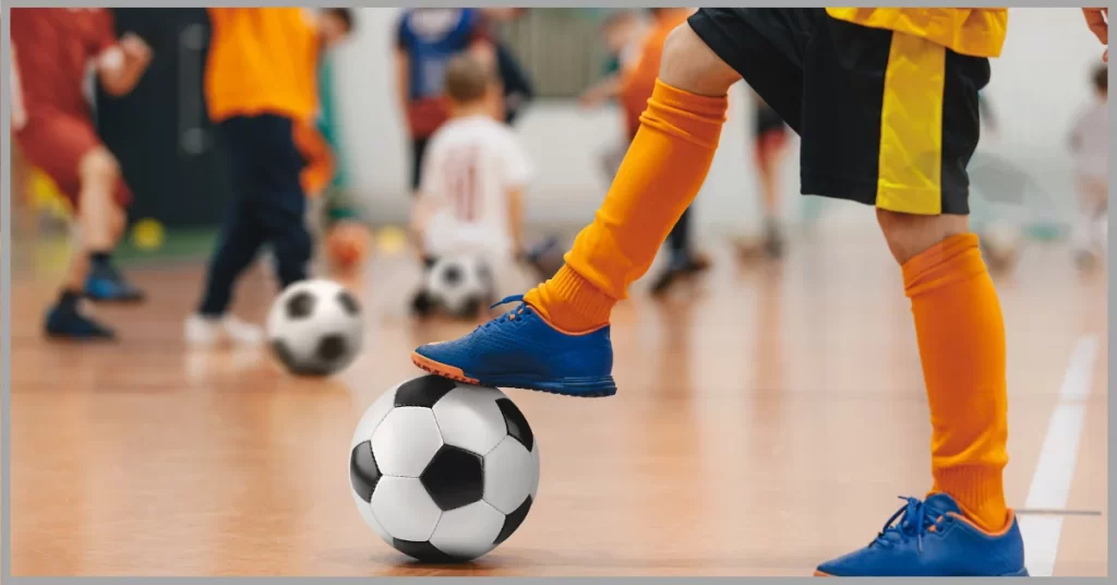 A child in a yellow and black soccer uniform is about to kick a soccer ball in an indoor gymnasium.