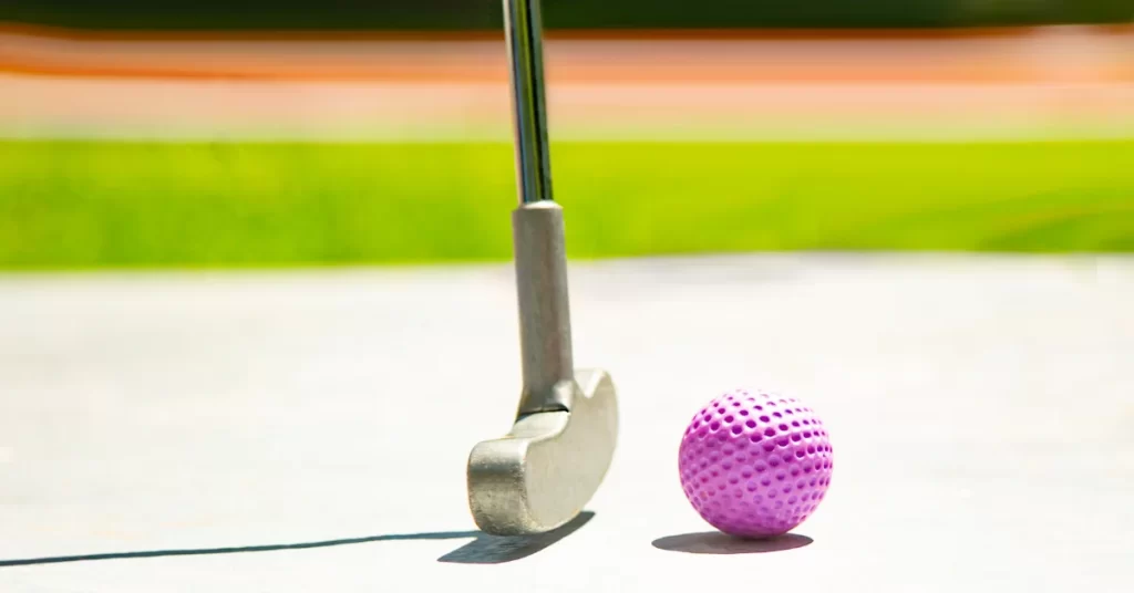 A close up of a pink golf ball and a putter on a mini golf course.