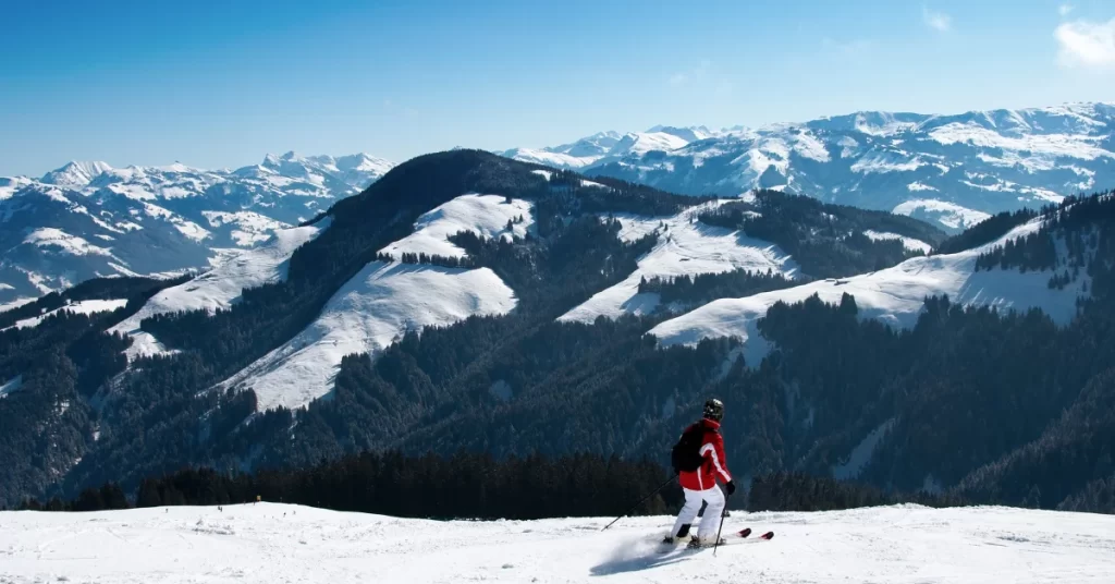 A skier in a red jacket and black pants glides down a snowy slope with a scenic view of the mountains and the sky in the background. (What Sports Are in Season)