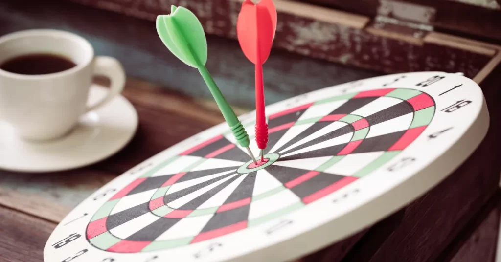 A photo of a dartboard with two darts on it, one green and one red, on a wooden table with a cup of coffee in the background. (Best Outdoor Dartboards)