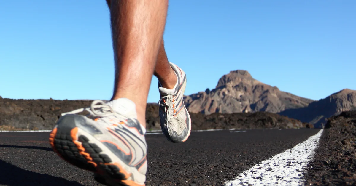 A person’s legs and feet, wearing sneakers, running on a paved road with a white line marking, surrounded by rocky terrain and mountains under a clear blue sky. (Cross-Training Shoes with Arch Support)