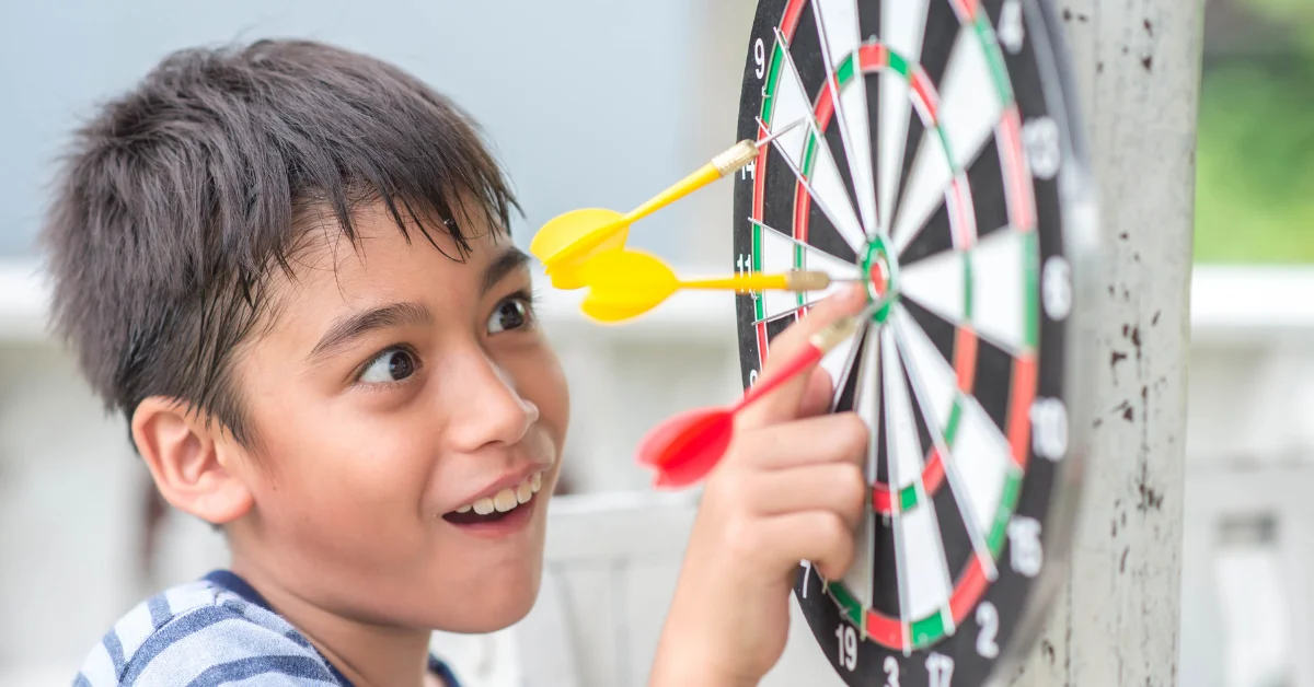 A person with their face blurred is aiming a dart at a dartboard attached to a tree.