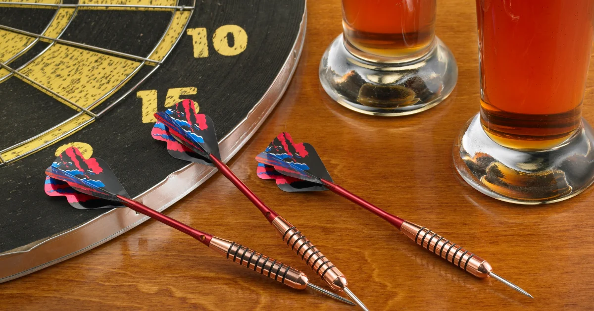 Two darts with red and blue flights are lying on a wooden surface next to a dartboard and two glasses of amber beer.